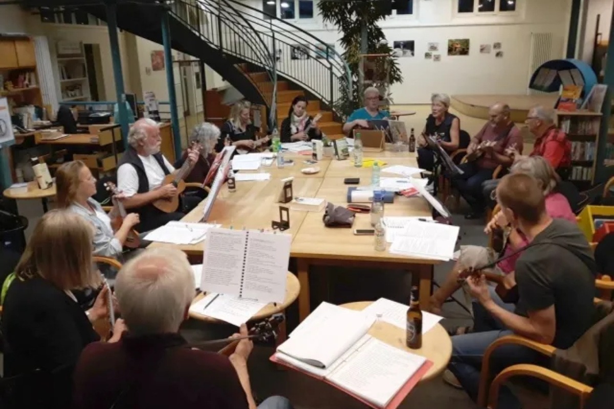 Foto mit einer Gruppe von Menschen, die Ukulele an einem runden Tisch mit Partituren spielen